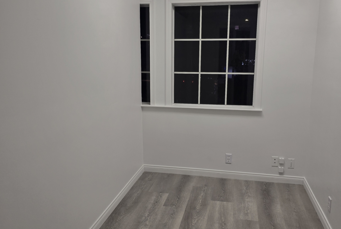 Bright empty salon room with large window, white walls, and gray wood flooring.