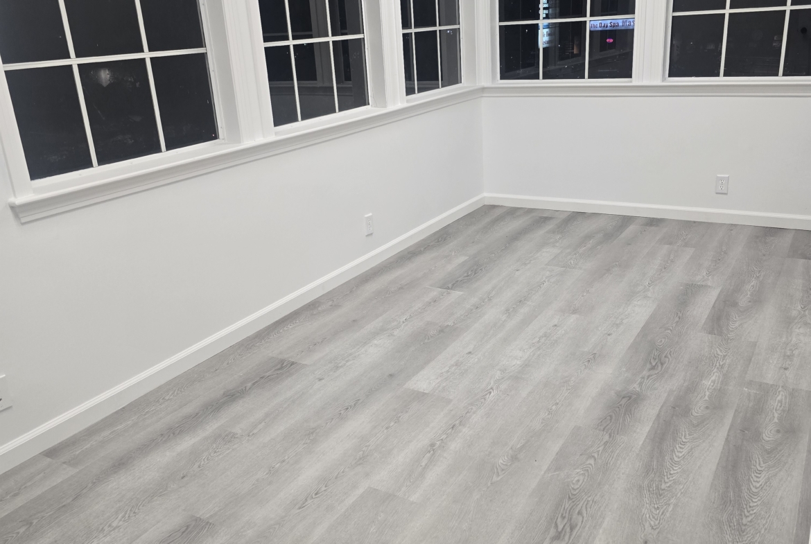 Bright salon room with multiple windows, white walls, and gray wood-look flooring, ideal for rental.