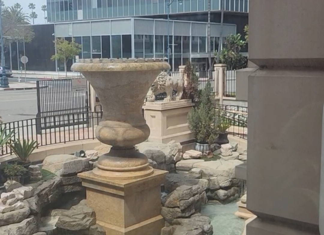 Exterior view of a sleek glass commercial building with indoor-outdoor garden featuring sculptures and water fountain.