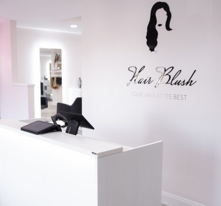 Modern hair salon reception desk with tech setup, styled wall logo, and welcoming ambiance for hair styling services.