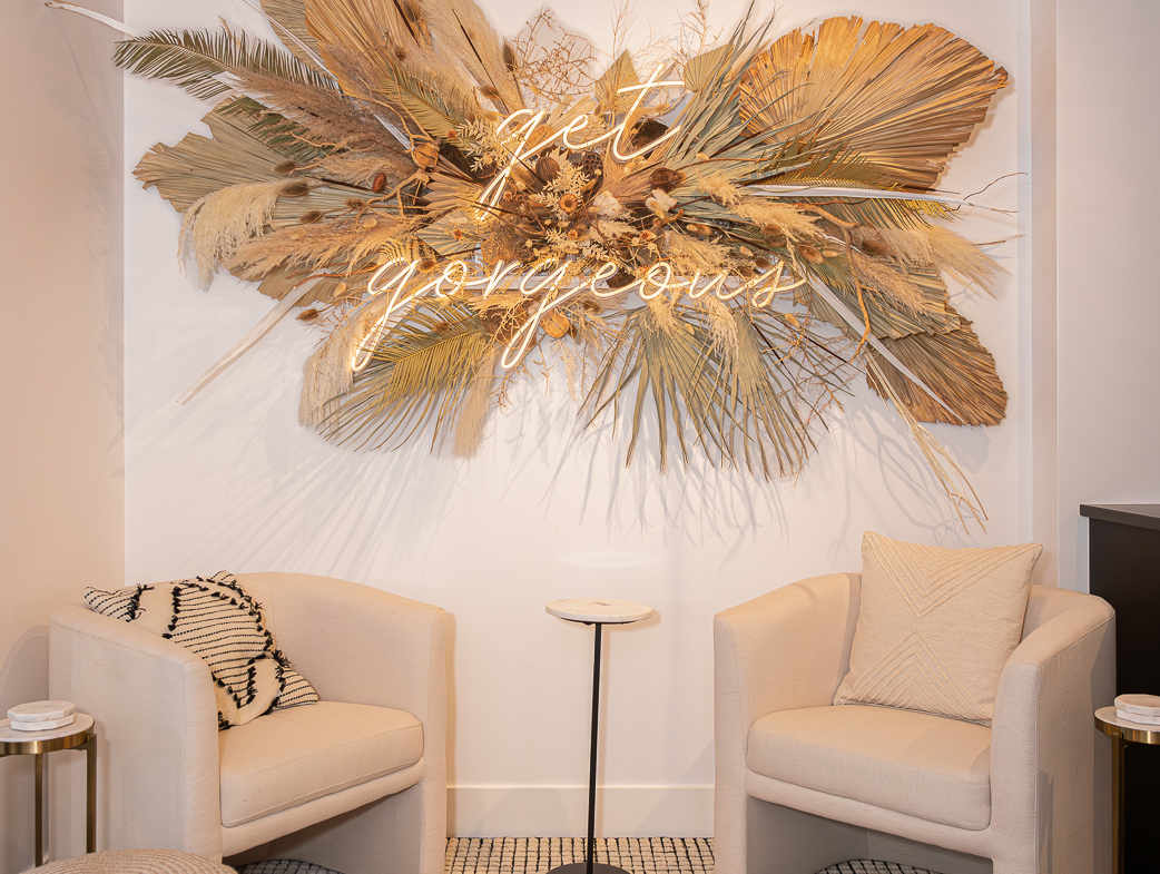 Dried floral wall art with neon "get gorgeous" sign, salon seating for a luxurious client experience.