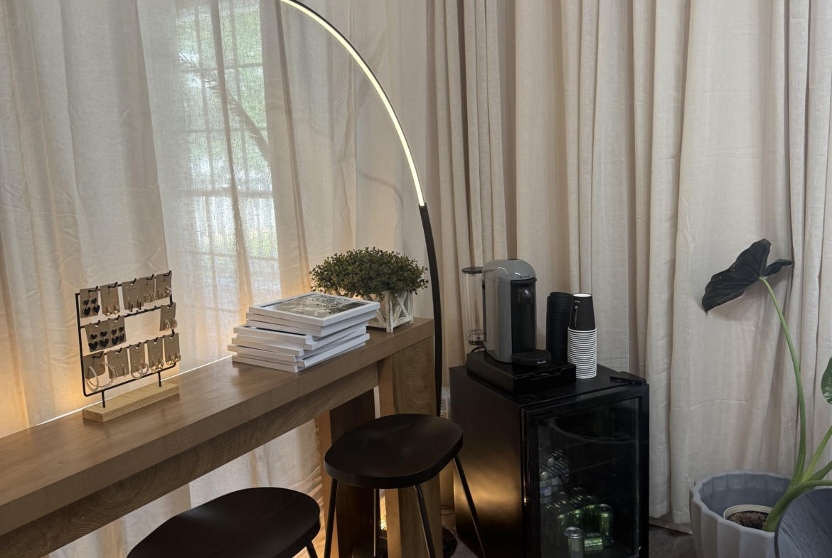 Alt: Salon lounge with black stools, coffee station, books, and indoor plant, modern decor.