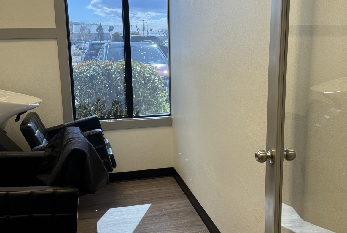 Bright salon interior featuring a styling chair near a large window with natural sunlight and parking view.