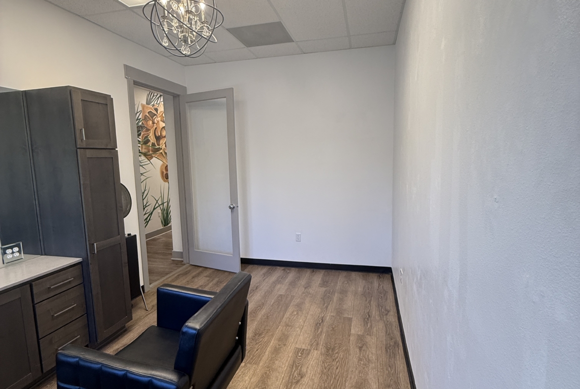 Bright, empty salon booth with styling chair, wood flooring, and ceiling light fixtures for rent.