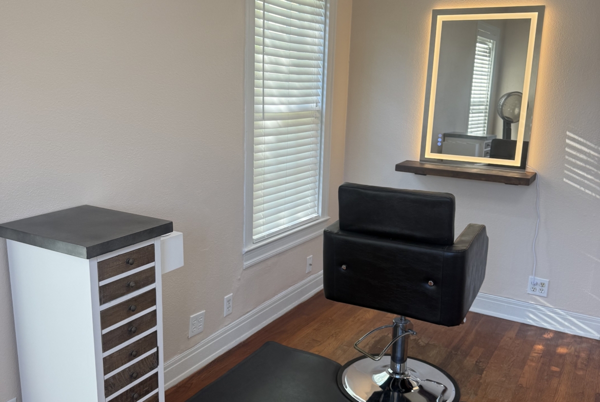 Comfortable salon styling area with black chair, large mirror, and natural light.