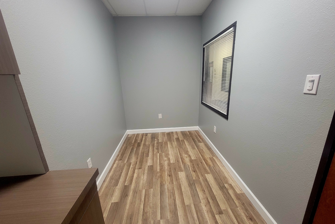 Bright, clean small room with neutral gray walls, wooden flooring, and window blinds, ideal for salon rental or beauty studio.