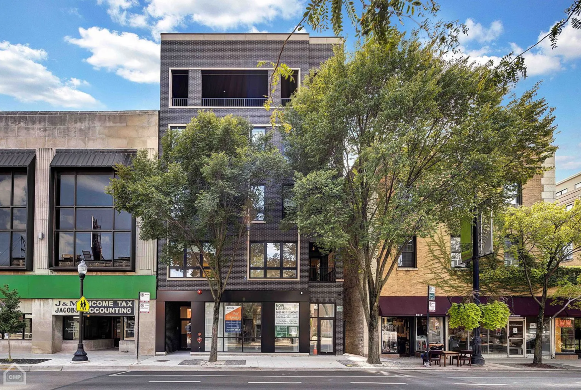 A contemporary city building with retail shops on the ground floor and residential units above, featuring glass windows and surrounding greenery.