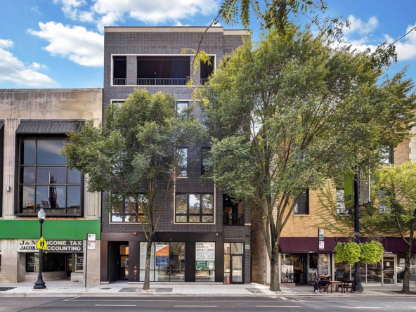 A contemporary city building with retail shops on the ground floor and residential units above, featuring glass windows and surrounding greenery.