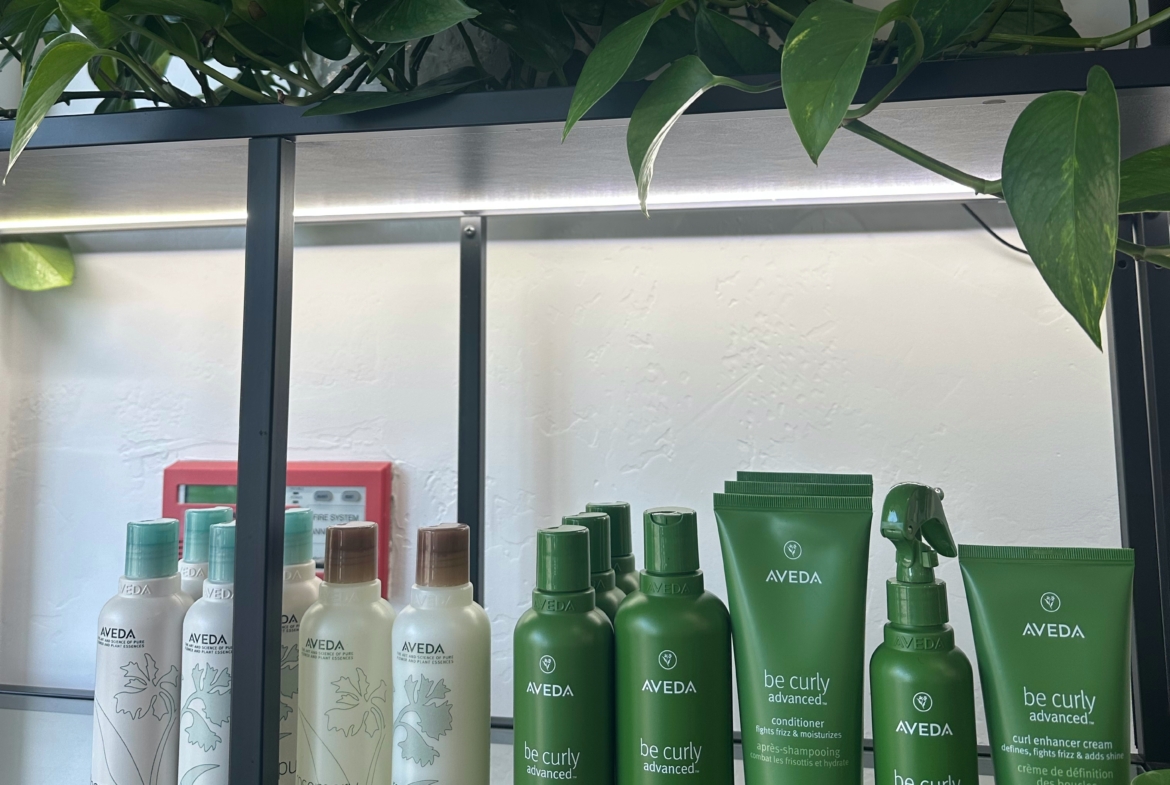 Aveda hair care products displayed on a modern shelf in a salon setting, featuring shampoos, conditioners, and styling sprays.