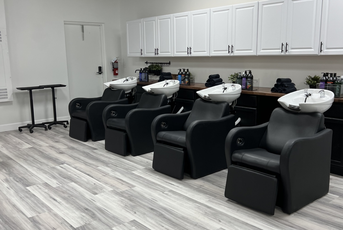 Comfortable salon wash area featuring black chairs, white sinks, and organized salon supplies.