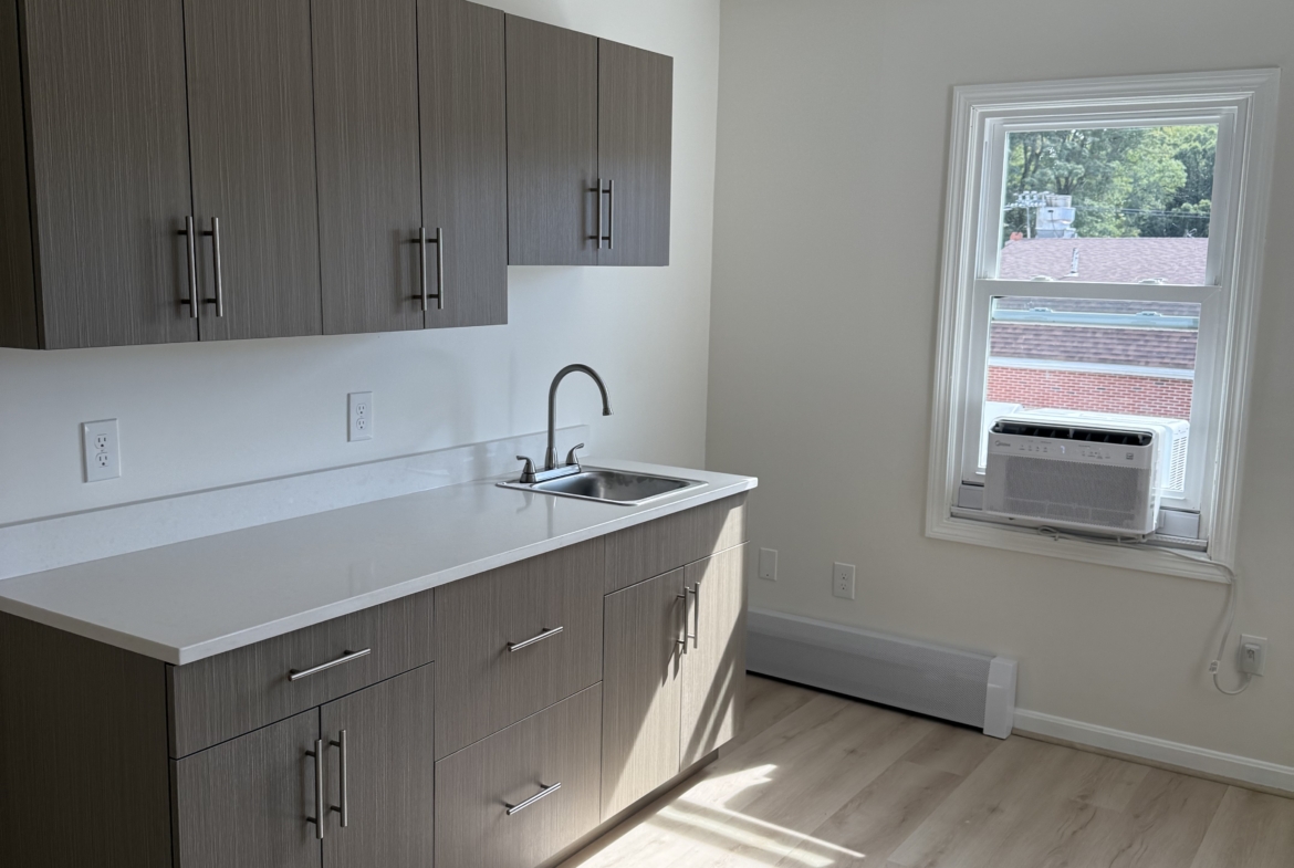 Bright rental salon kitchen with wood cabinetry, window air conditioning, and hardwood floor.