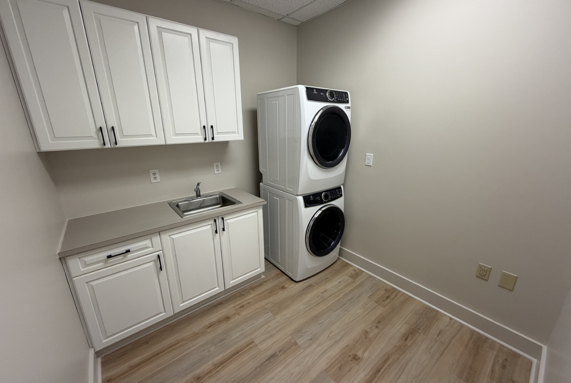 Clean laundry room featuring stacked washer and dryer, white cabinets, and a utility sink for salon staff convenience.