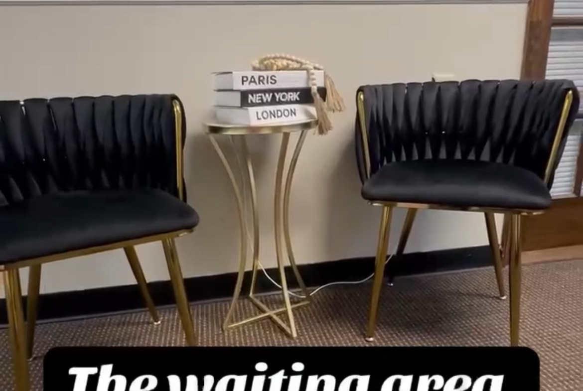 A chic salon waiting area with two black upholstered chairs with gold accents, a small decorative table, and travel books for clients.
