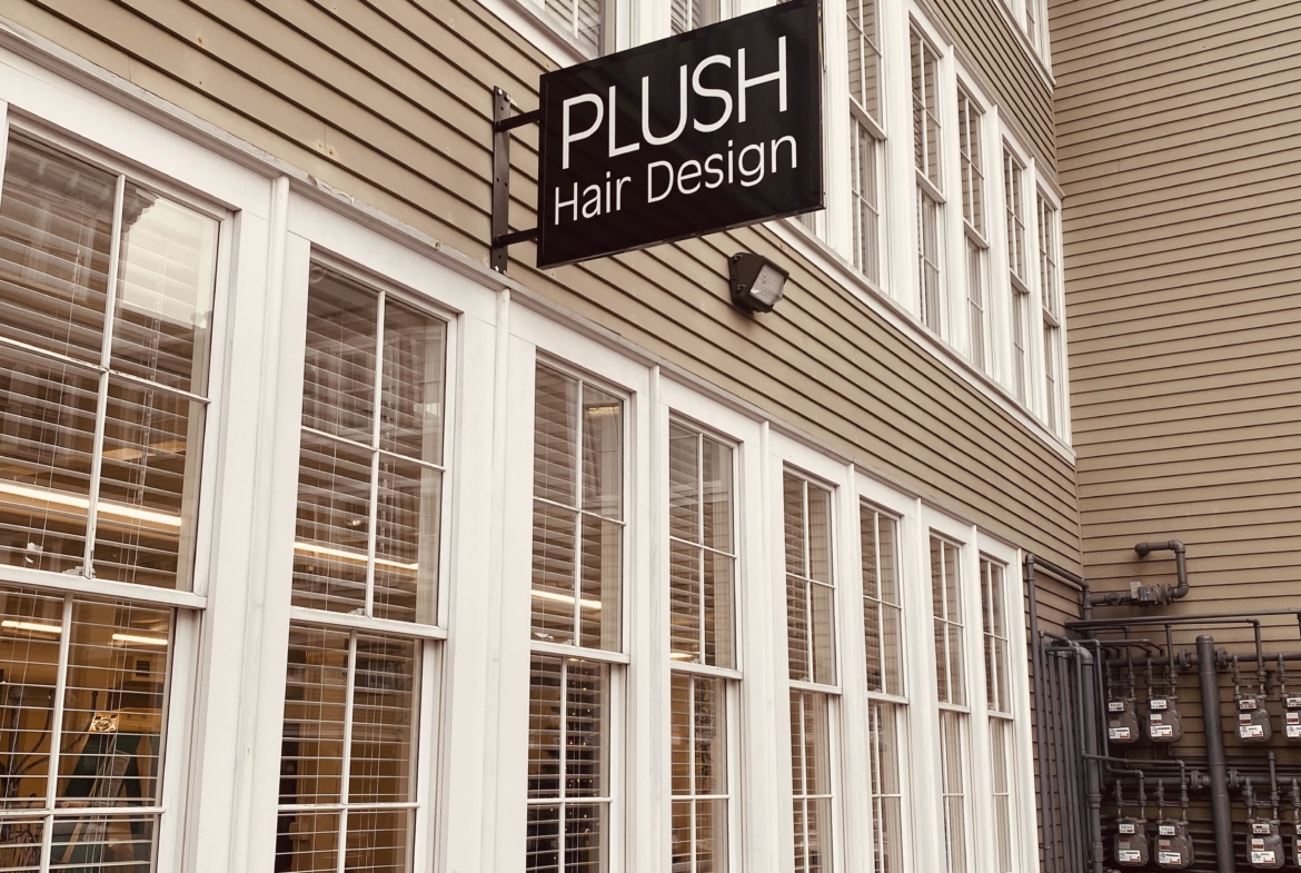 Exterior of hair salon with "PLUSH Hair Design" sign on beige siding building, downtown urban scene.