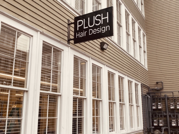 Exterior of hair salon with "PLUSH Hair Design" sign on beige siding building, downtown urban scene.