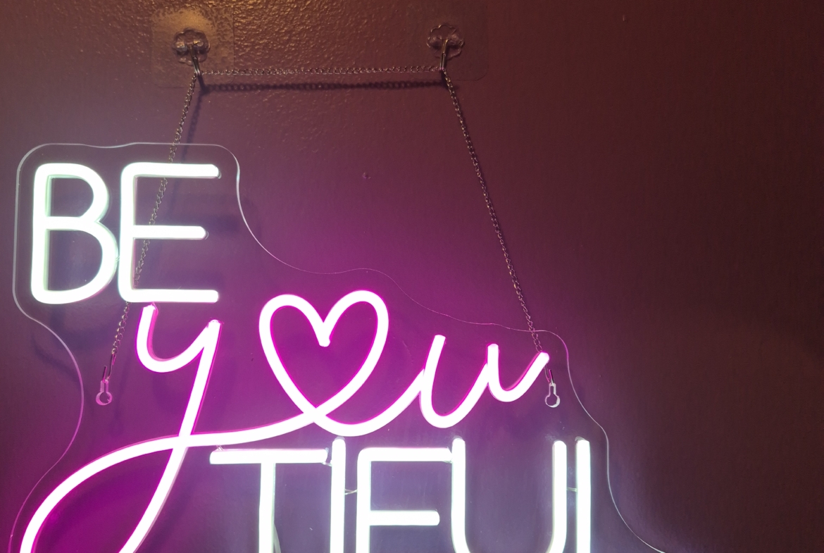 Neon sign reading "Be You Tiful" with pink and white colors on dark wall background.