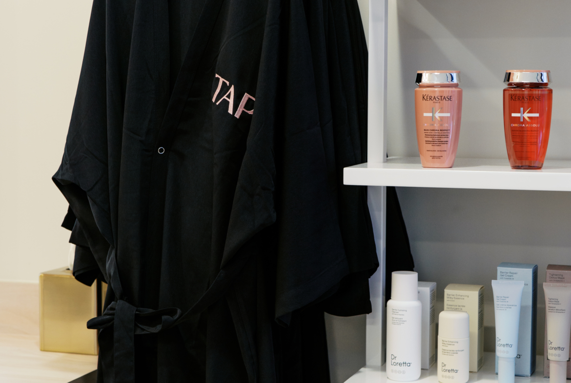 Close-up of black salon robe and hair care products on white shelves, featuring Kerastase and Dr. Loretta.
