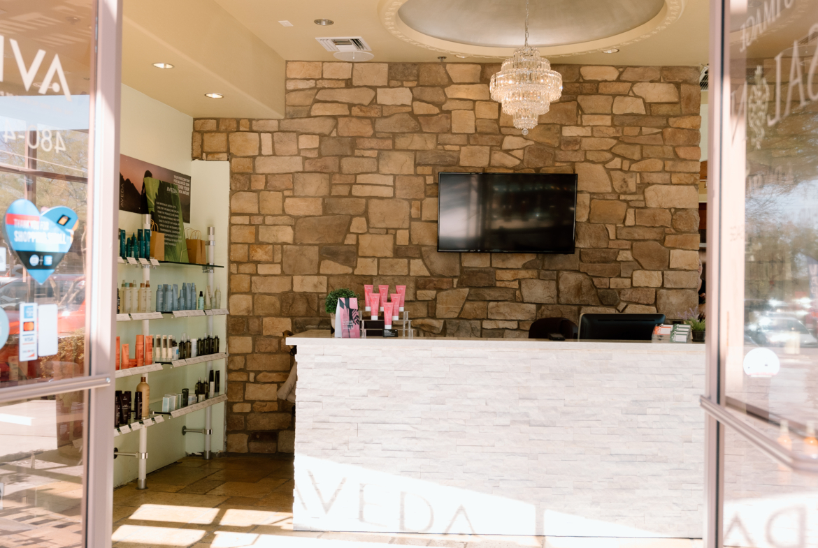 Interior view of a modern beauty salon reception with a stone accent wall, chandelier, and product displays.