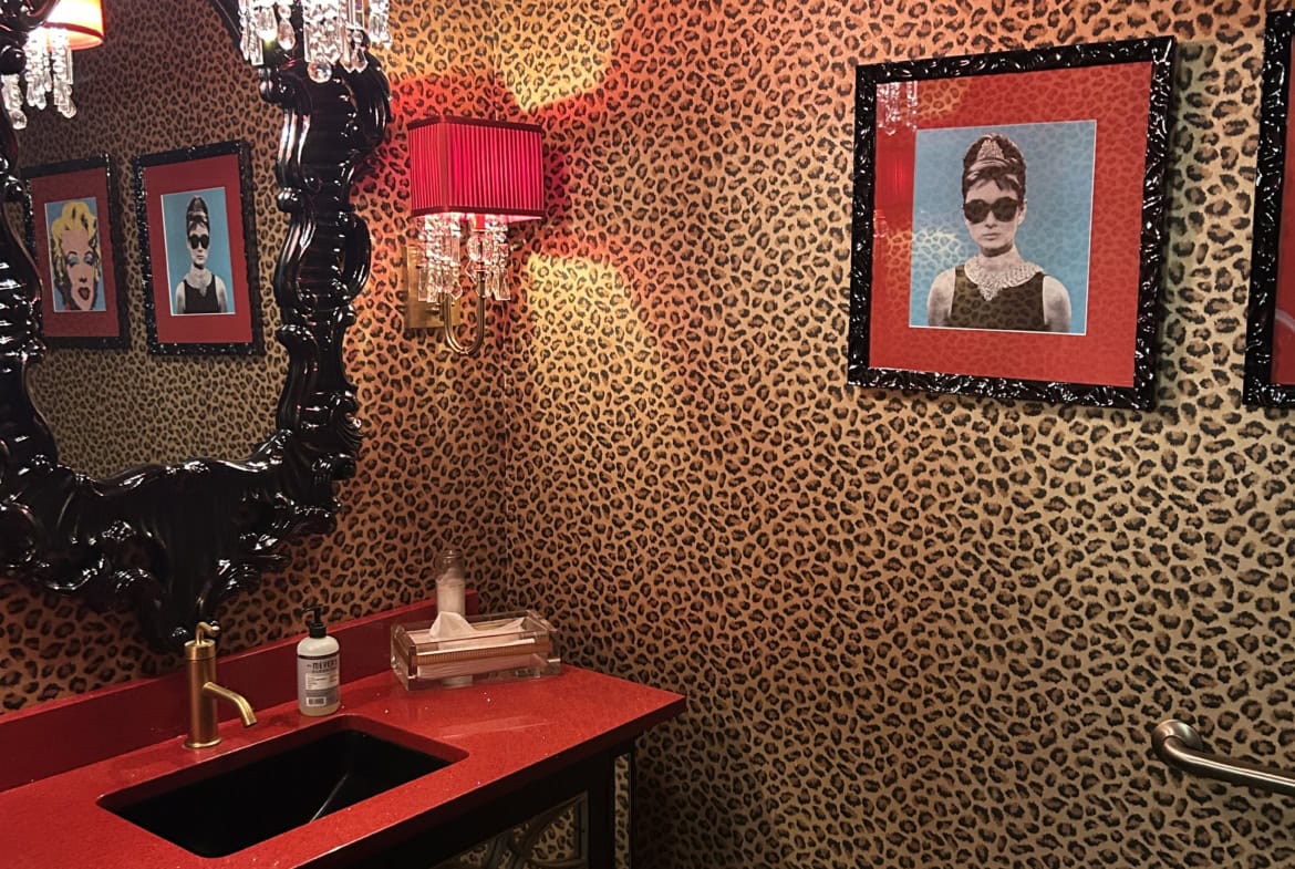 Luxurious salon interior with leopard print wallpaper, black ornate mirror, red sink, and framed artwork of stylish women.