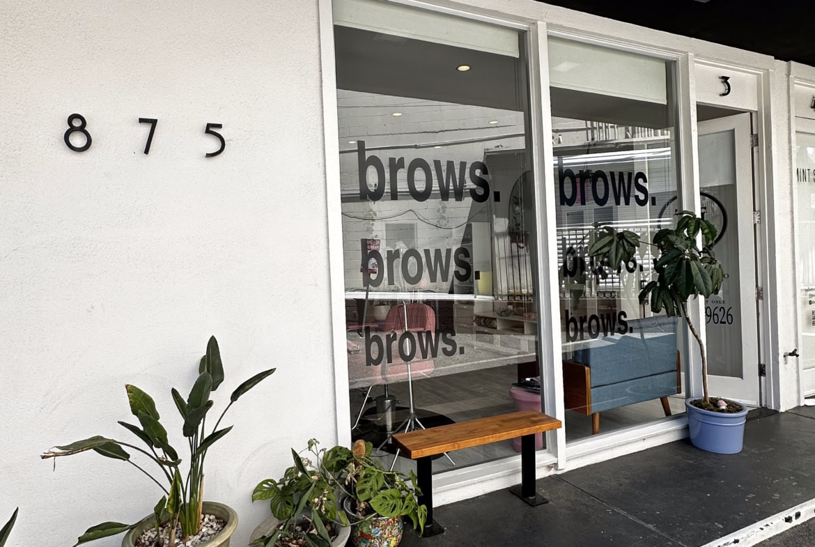 Modern salon storefront with large glass window displaying "brows" signage, outdoor plants, and inviting entryway.