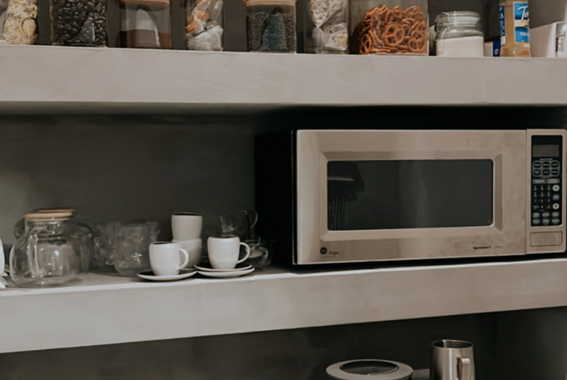 Microwave and coffee machine in a salon kitchenette, featuring organized shelves with jars and a sle.