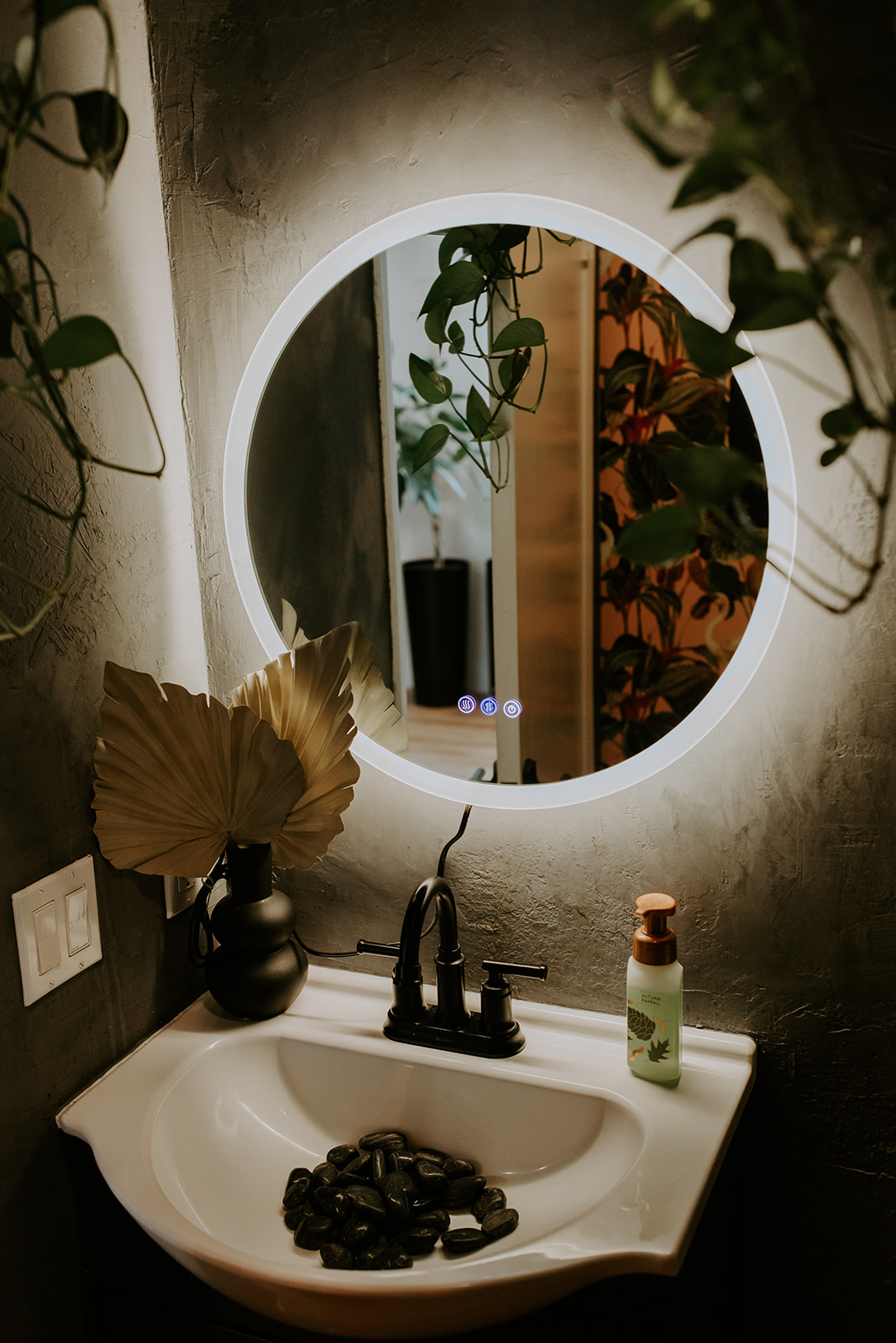 Round illuminated mirror above a bathroom sink with plants and decor, stylish and modern design.