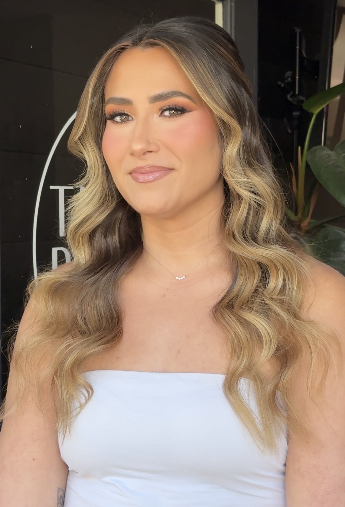 Beautiful woman with long, wavy blonde hair, makeup, and a white top, showcasing hair styling servic.