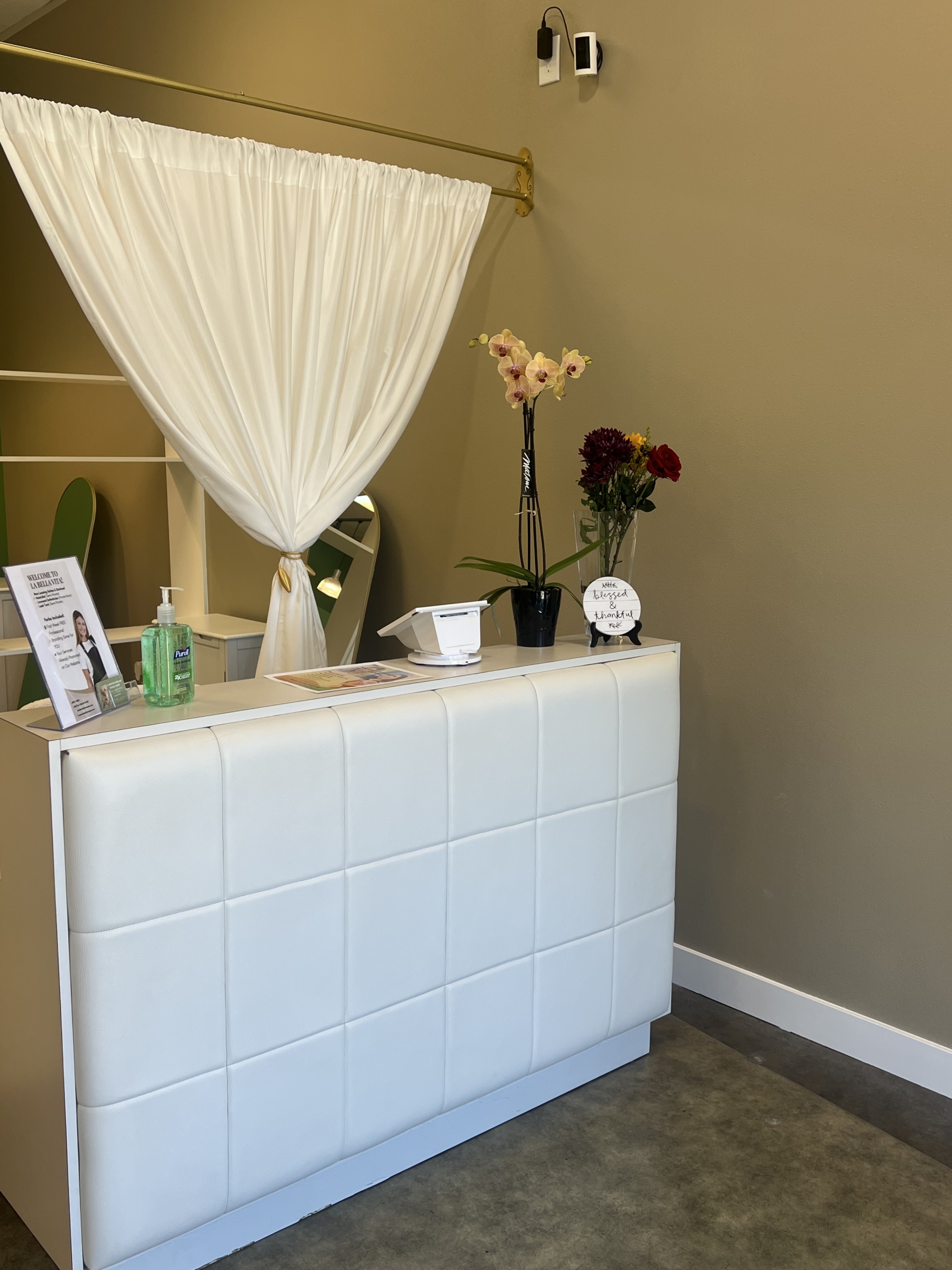 Modern salon reception area with white padded desk, floral arrangements, and privacy curtain.