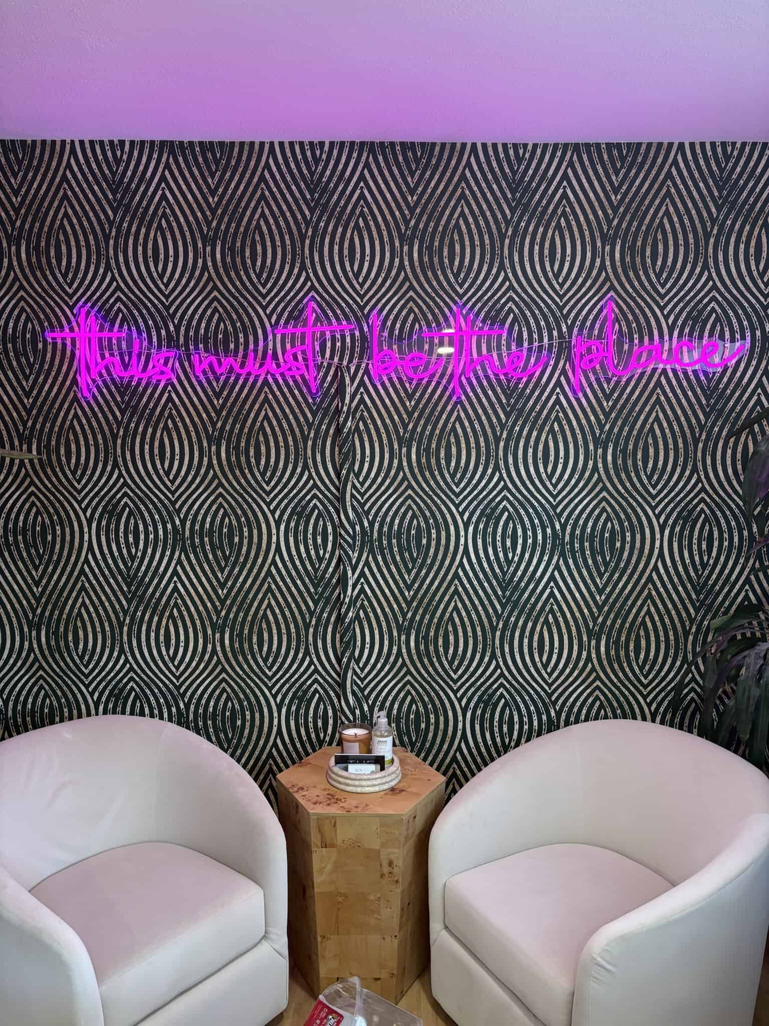 Neon sign reading "the wait here place" on stylish patterned wall in modern salon lounge. Comfortabl.